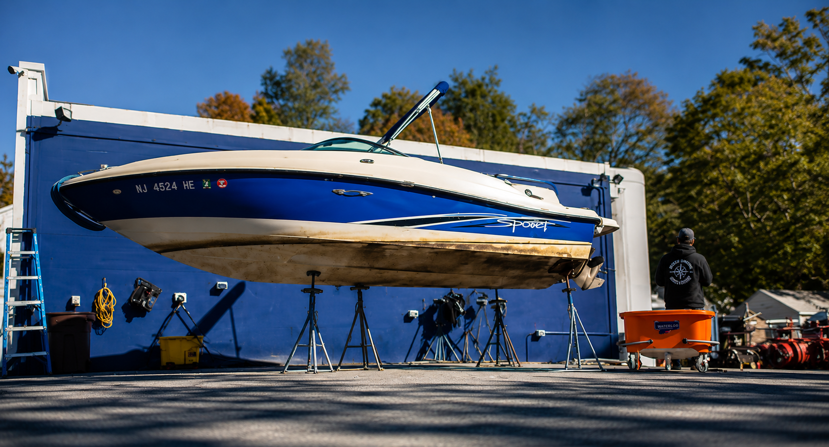 Blue-and-white boat lifted on a boat hoist, with people and equipment underneath outdoors