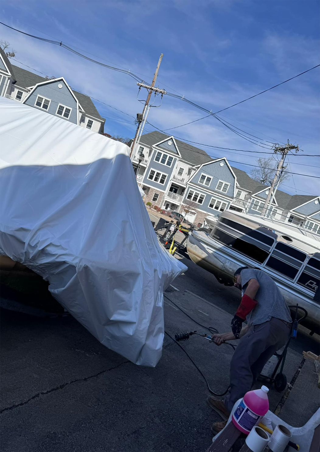 Covered boat on a street beside parked cars, with houses and a blue sky in the background.