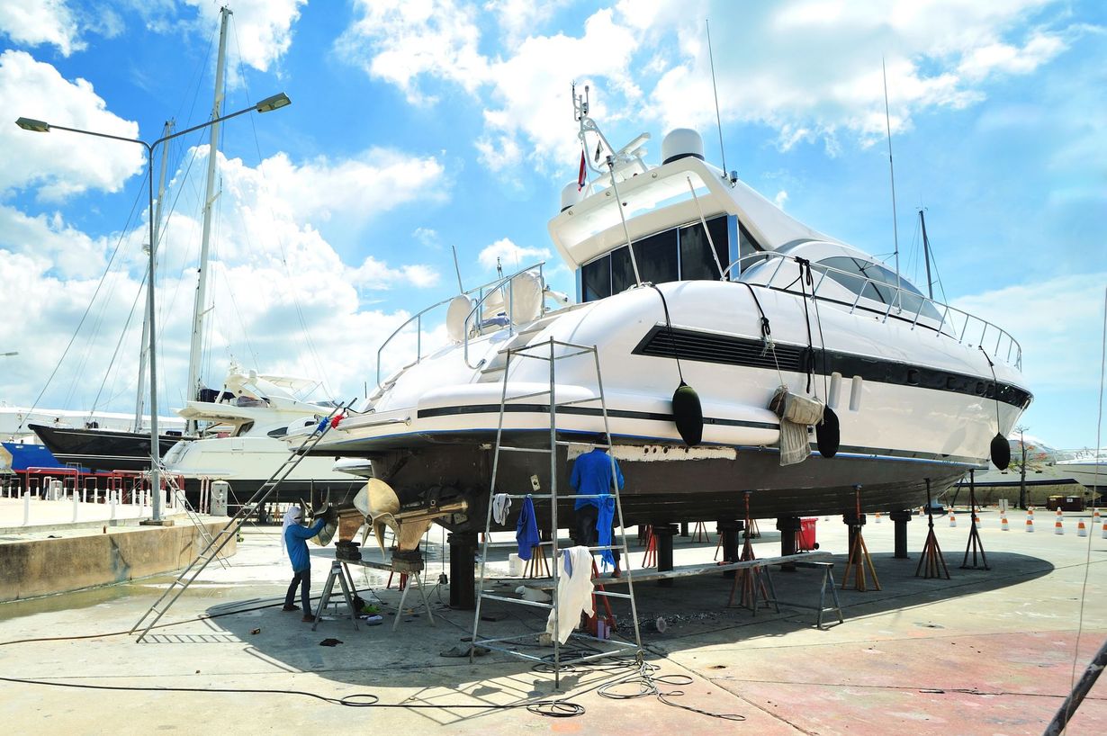 A white motor yacht on supports in a shipyard with technicians performing maintenance on its propellers.