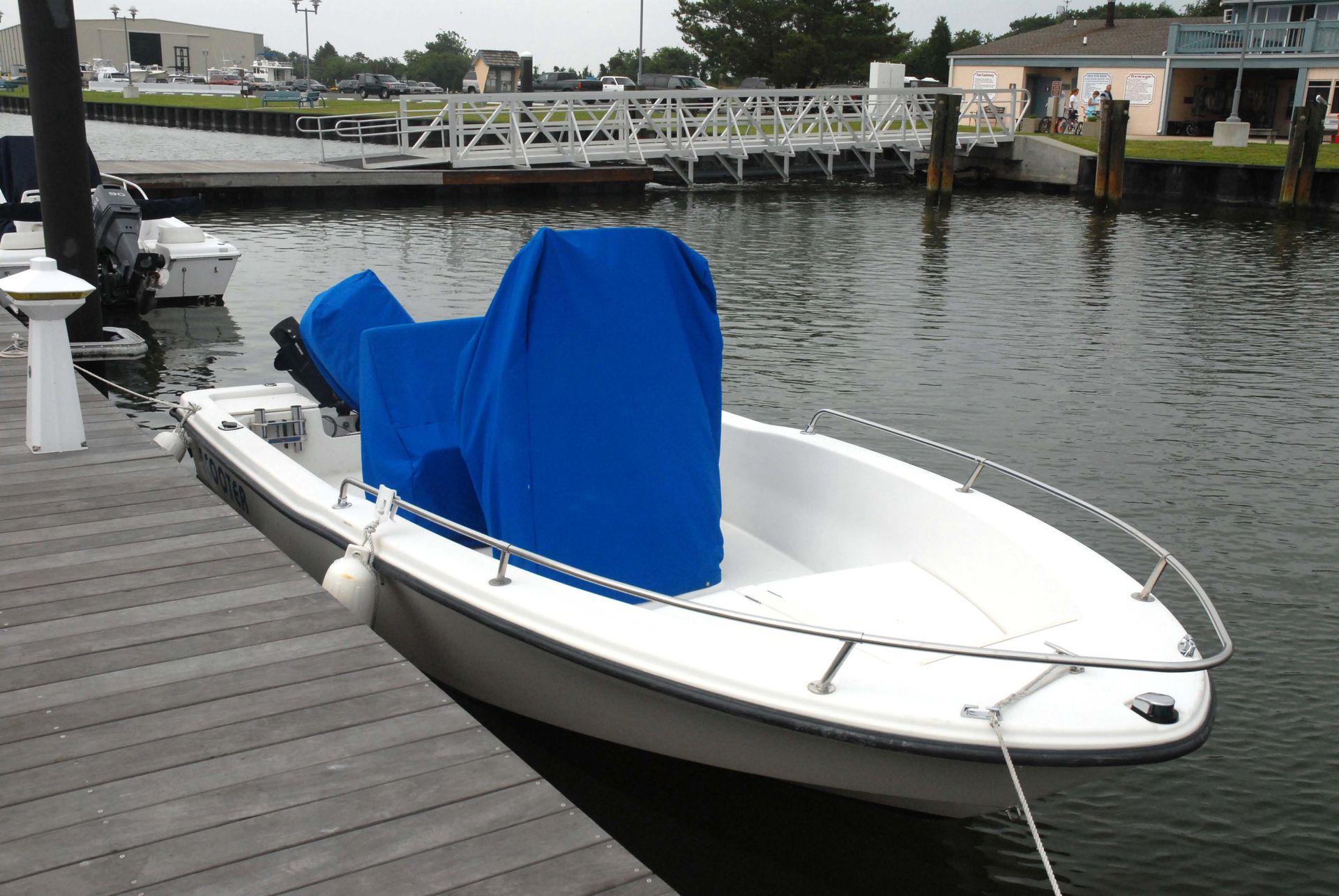 A small white motorboat docked at a wooden pier with a blue console cover, next to a marina and building.