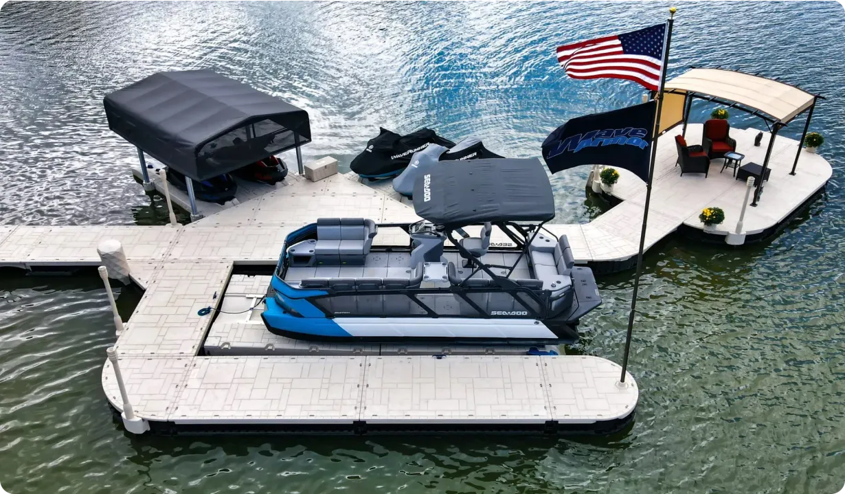 An aerial view of a boat dock with a docked blue and gray pontoon boat, a covered jet ski lift, and a seating area.