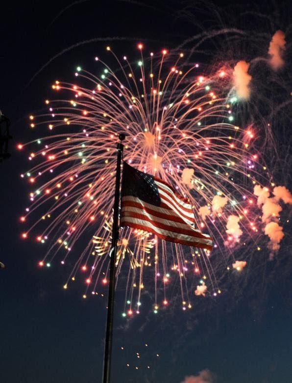 American flag on a pole silhouetted against colorful fireworks in a night sky