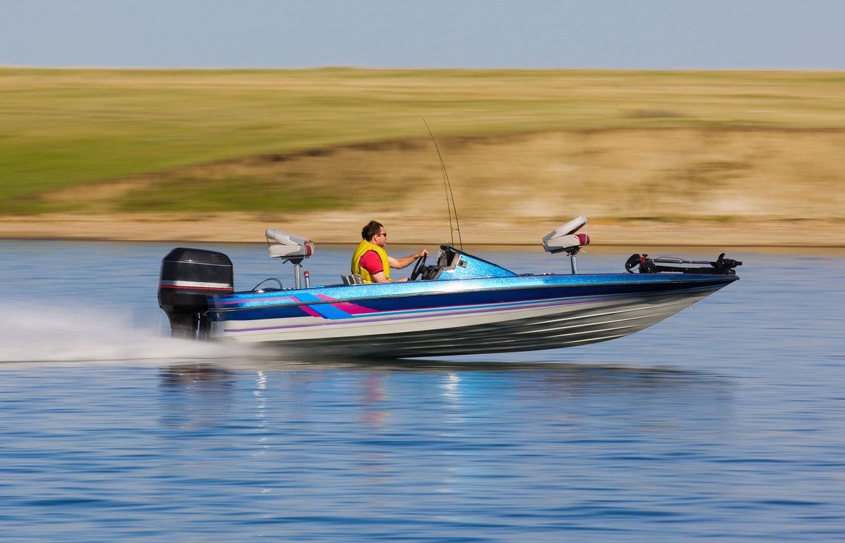 A person in a yellow life vest drives a blue and silver bass boat quickly across a calm lake with a grassy shore behind.