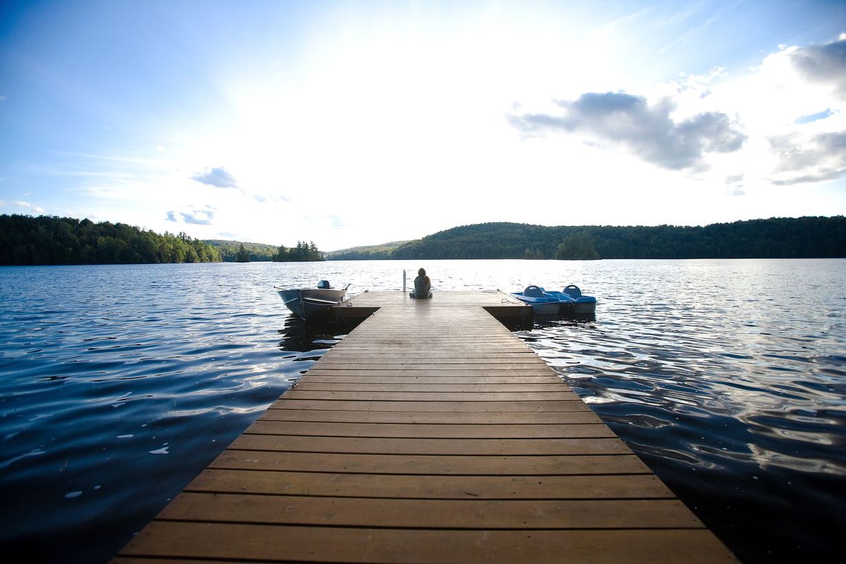 A person sits at the end of a wooden dock extending into a calm lake surrounded by forested hills under a bright sky.