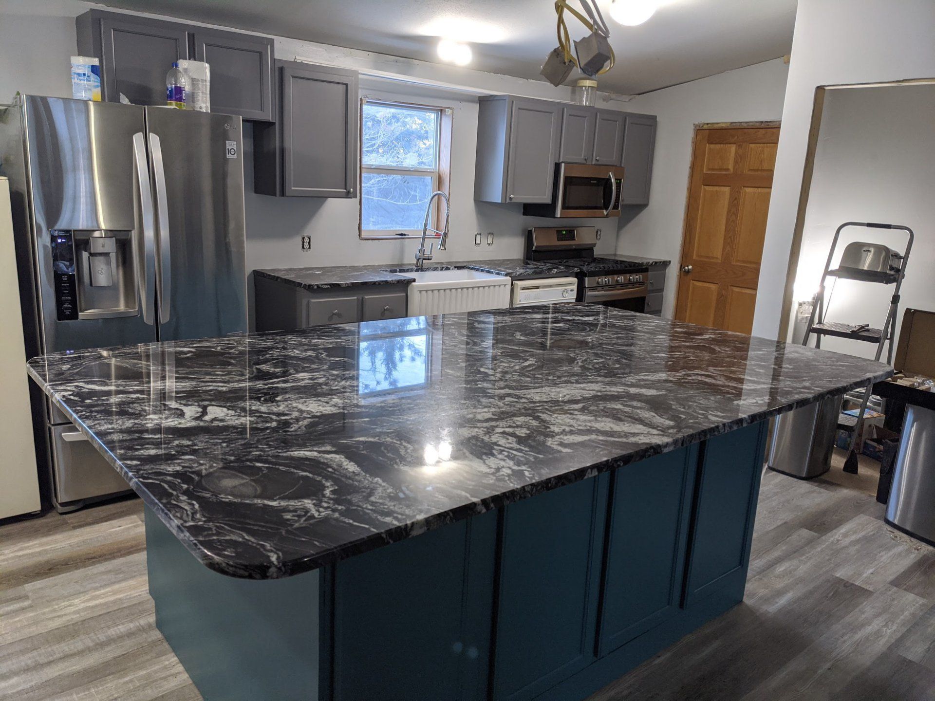 Kitchen with island; dark countertop, blue cabinets, stainless steel appliances, grey upper cabinets, and a farmhouse sink.