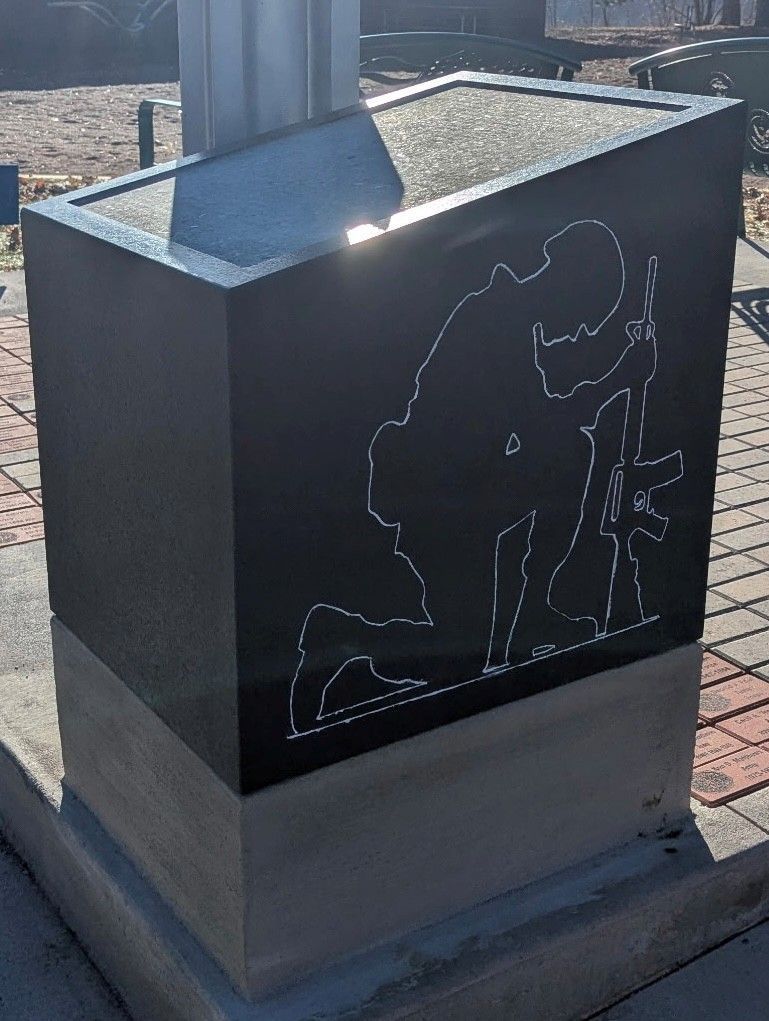 Black monument with a soldier kneeling, etched, atop a concrete base. Sunlight gleams on the angled surface.