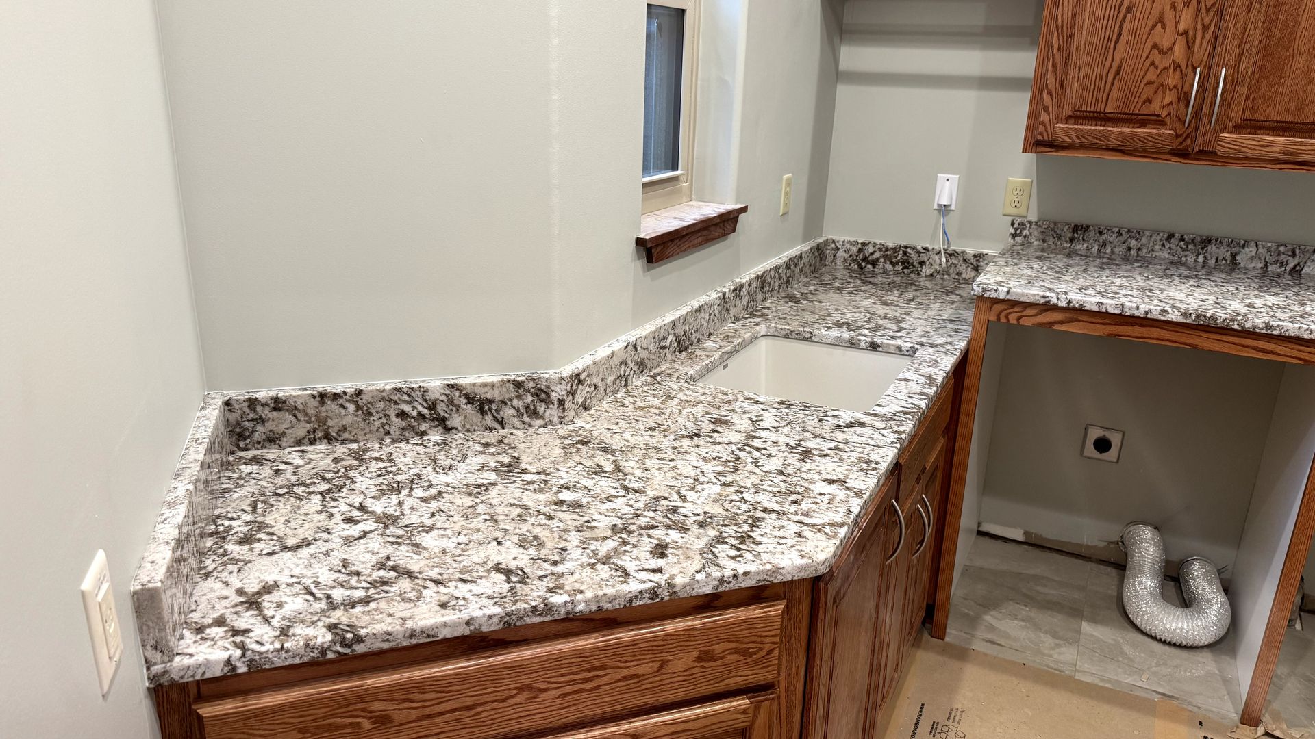 A kitchen counter with granite countertops and wood cabinets, near a window, with a sink.