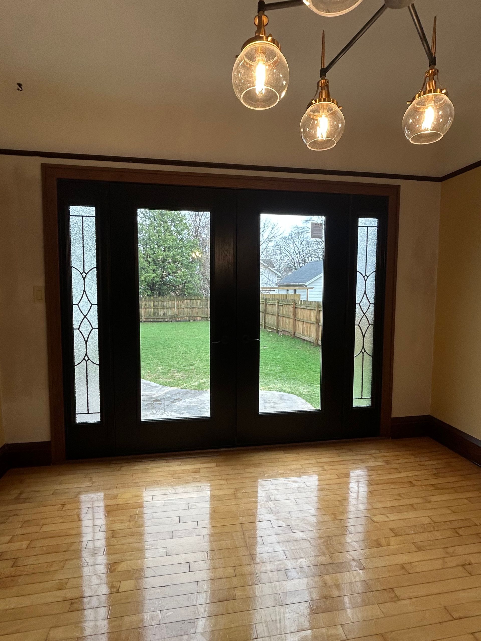 Empty room with wood floor, black French doors, and a hanging three-light fixture overlooking a green yard