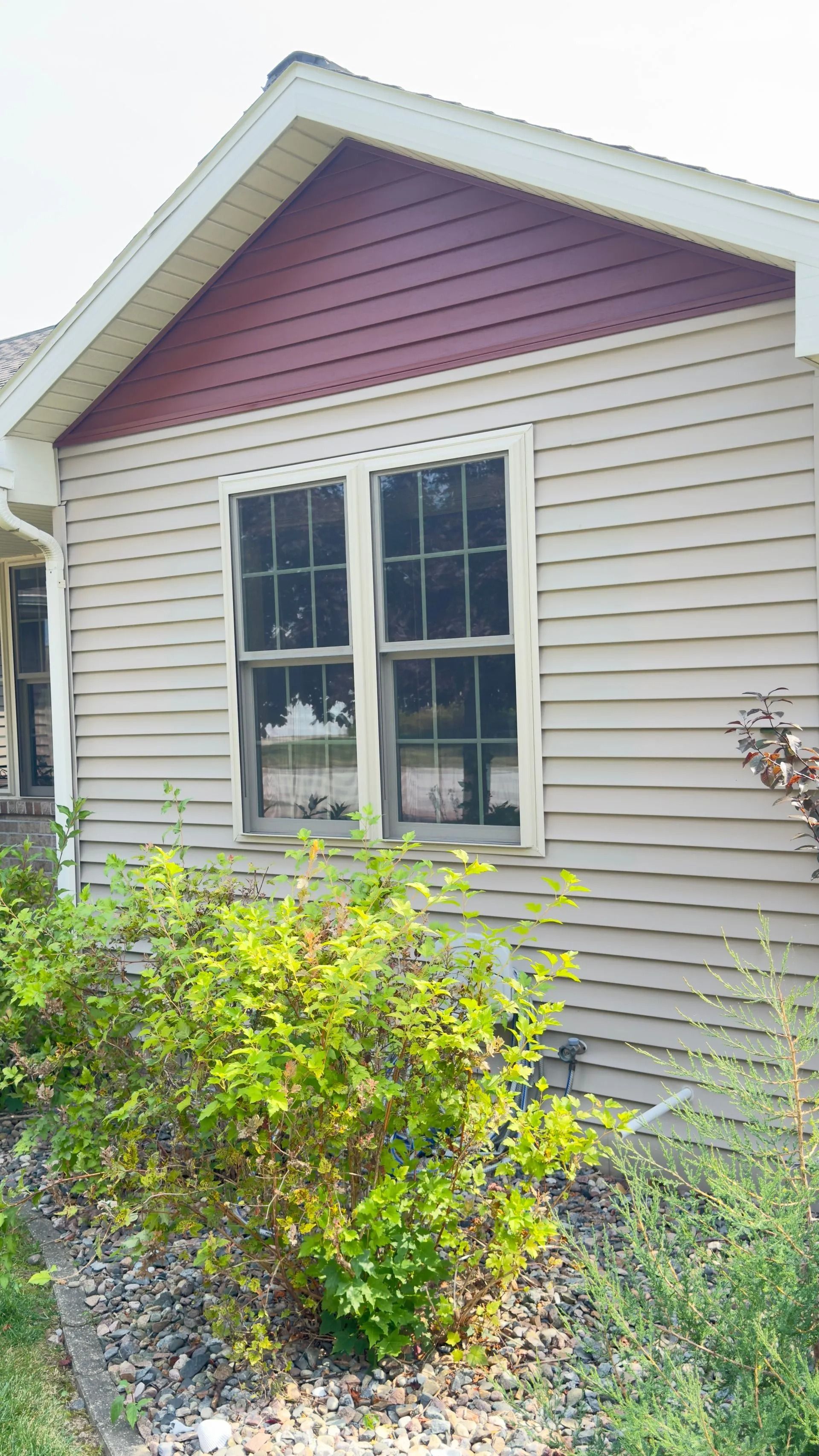 Tan house exterior with window and red gable. Green bushes in front.