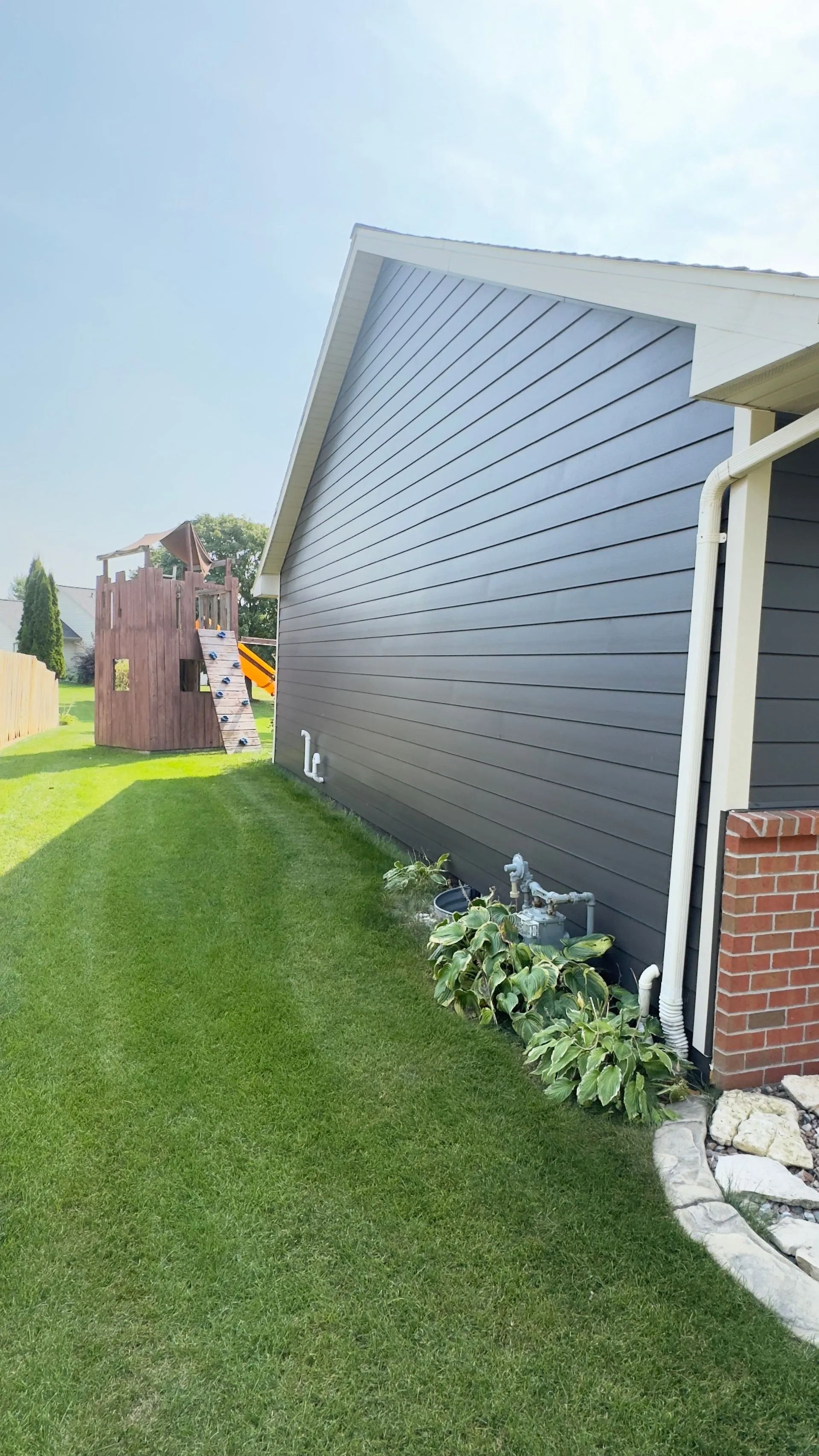 Side view of a house with dark gray siding, green lawn, and a wooden playset in the background.