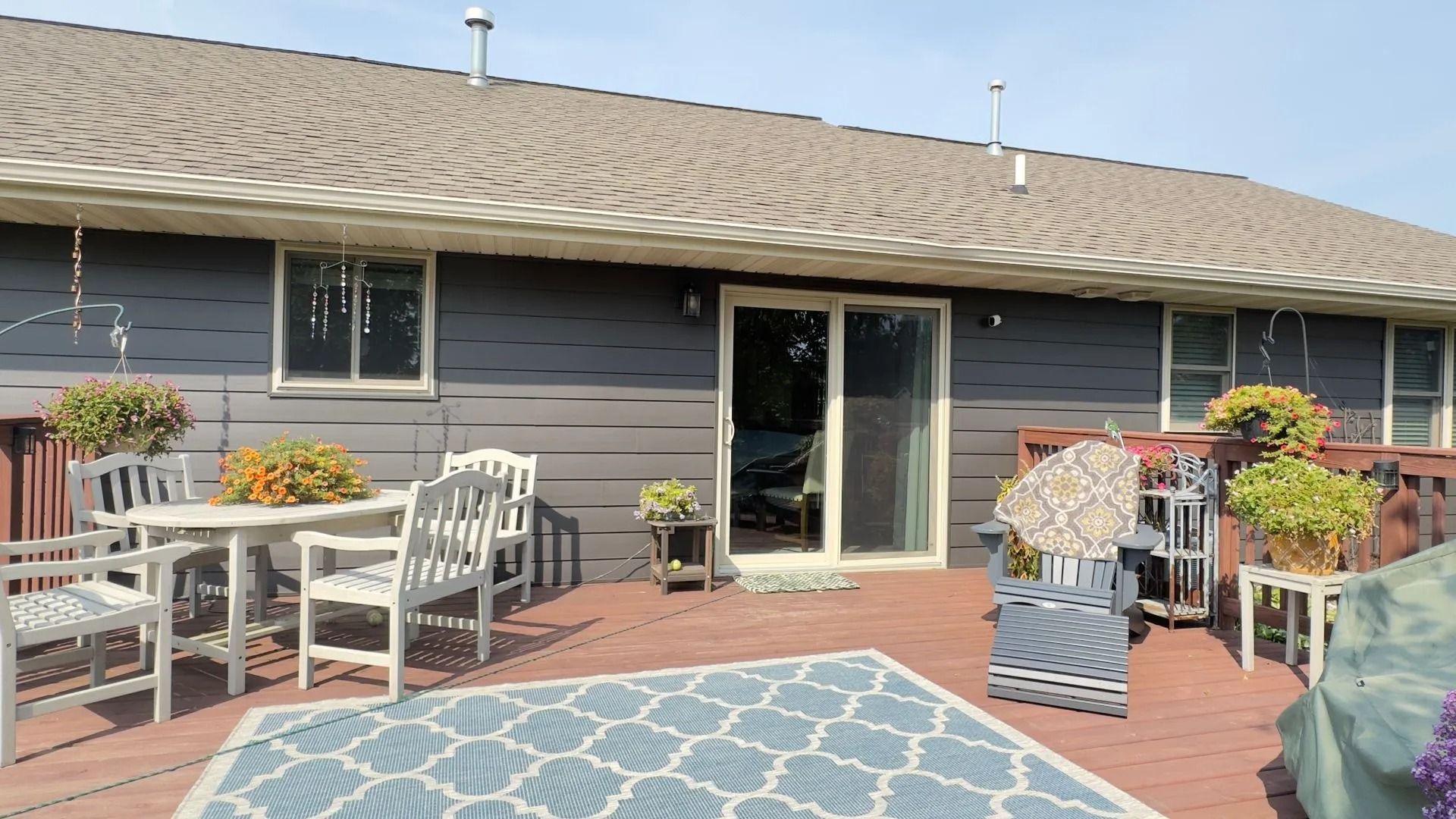 Wooden deck with white chairs and table, sliding glass door, and a gray house exterior.