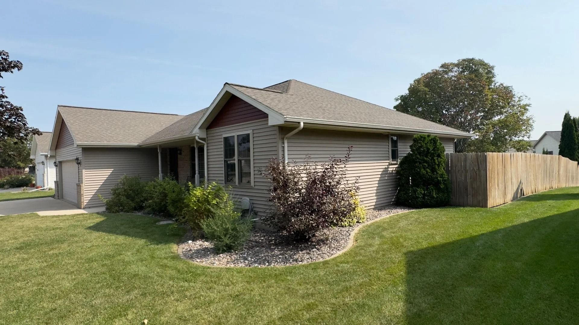 Tan house with a brown roof and wooden fence, with bushes in the front yard under a blue sky.
