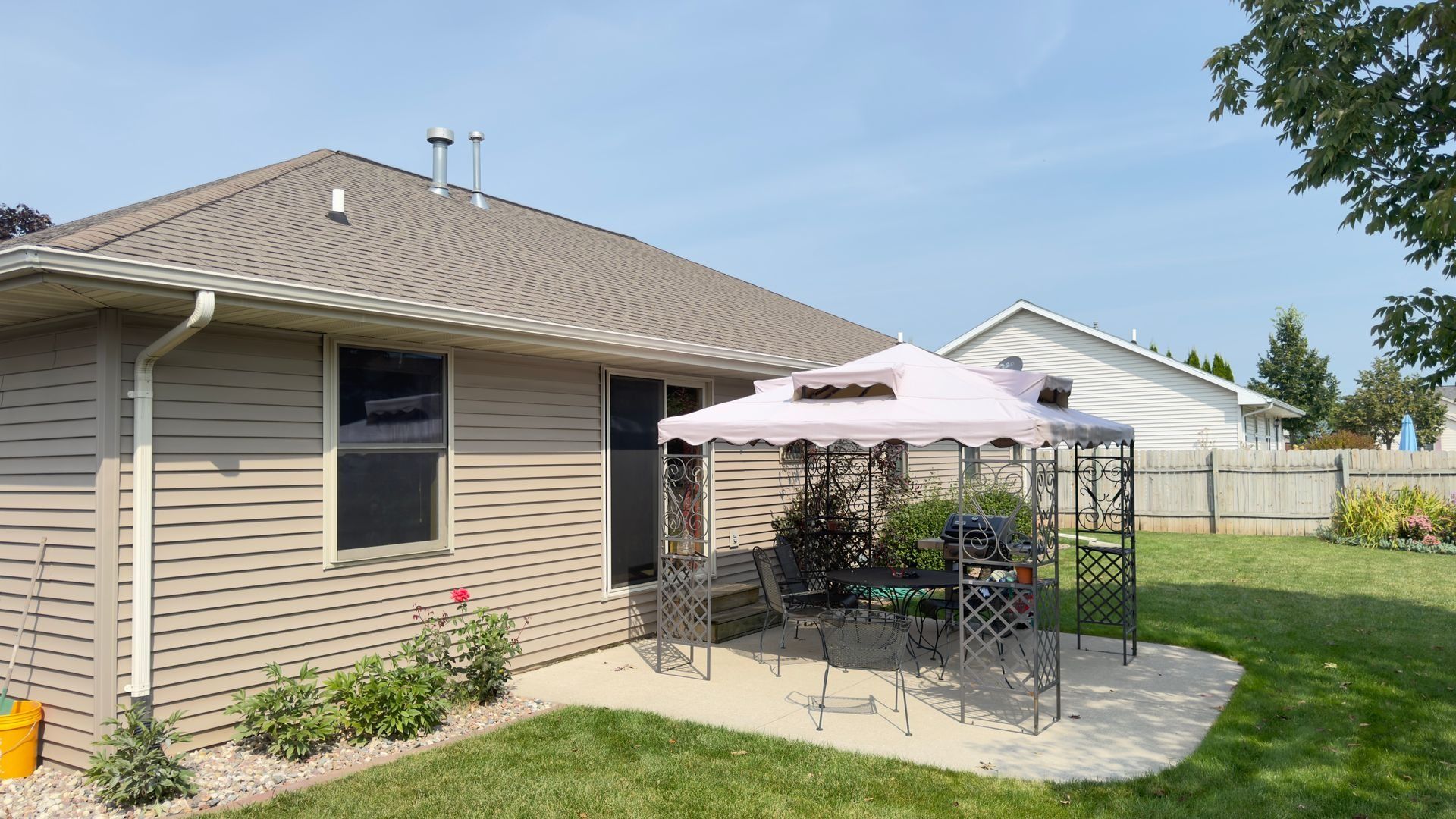 Backyard with beige house, patio, gazebo, and green grass on a sunny day.