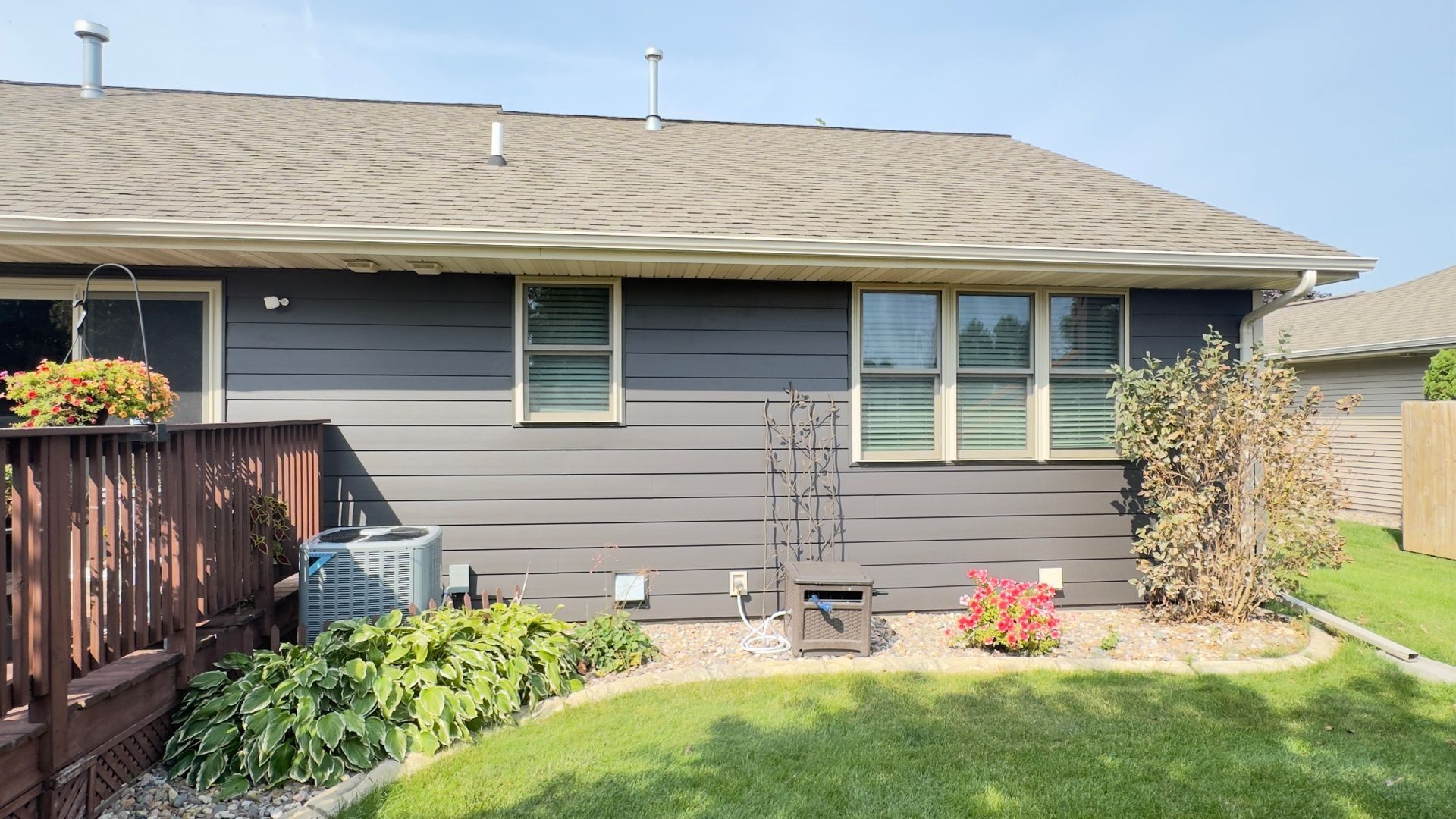 Backyard view of a house with gray siding, windows, and a brown deck, with a small garden and lawn.