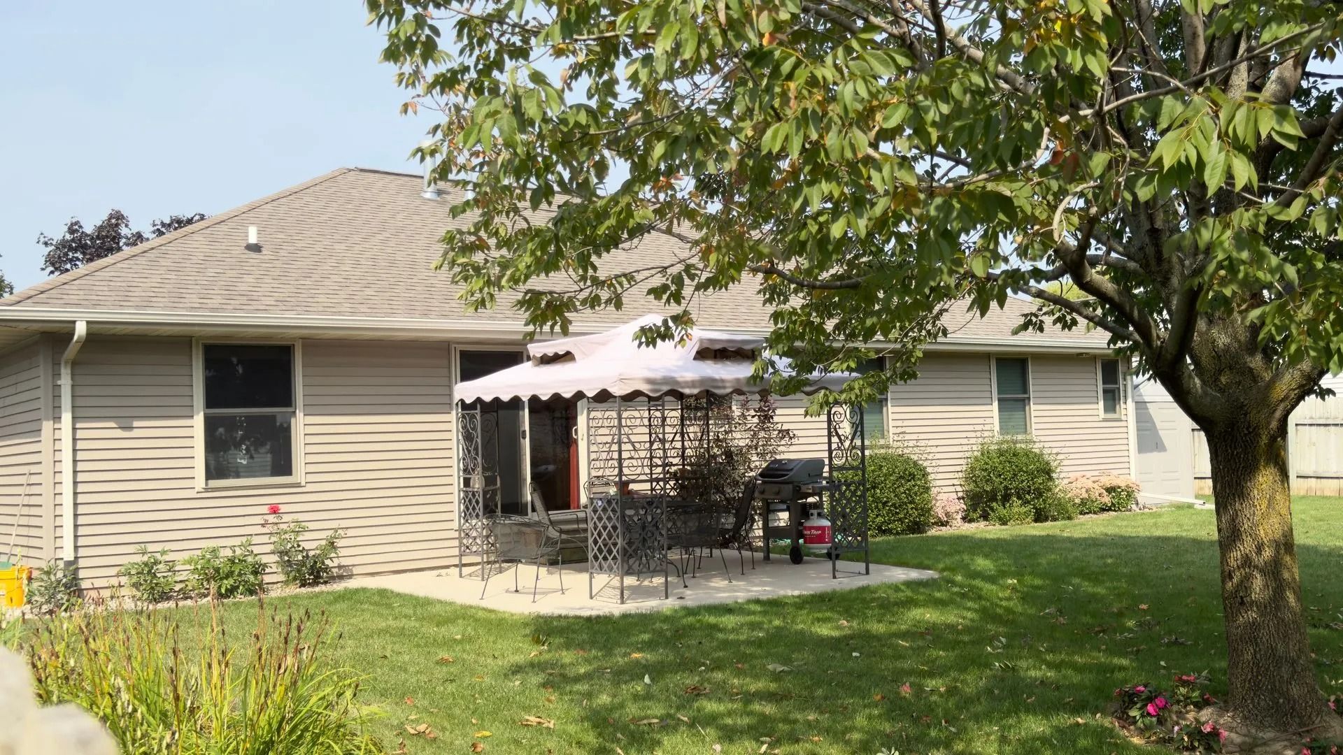 Backyard patio with table, chairs, grill, and gazebo, next to house with green lawn and tree.