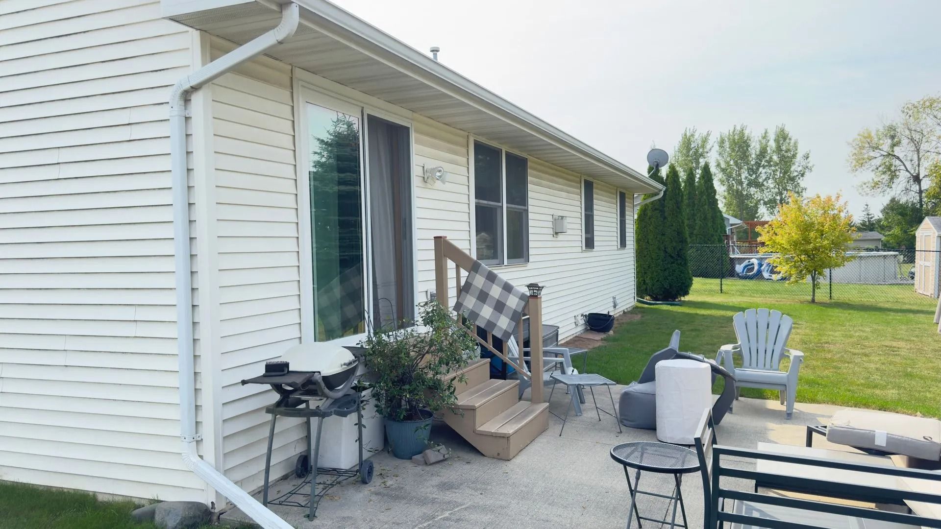 Backyard patio with sliding door, grill, wooden steps, and chairs on a sunny day.
