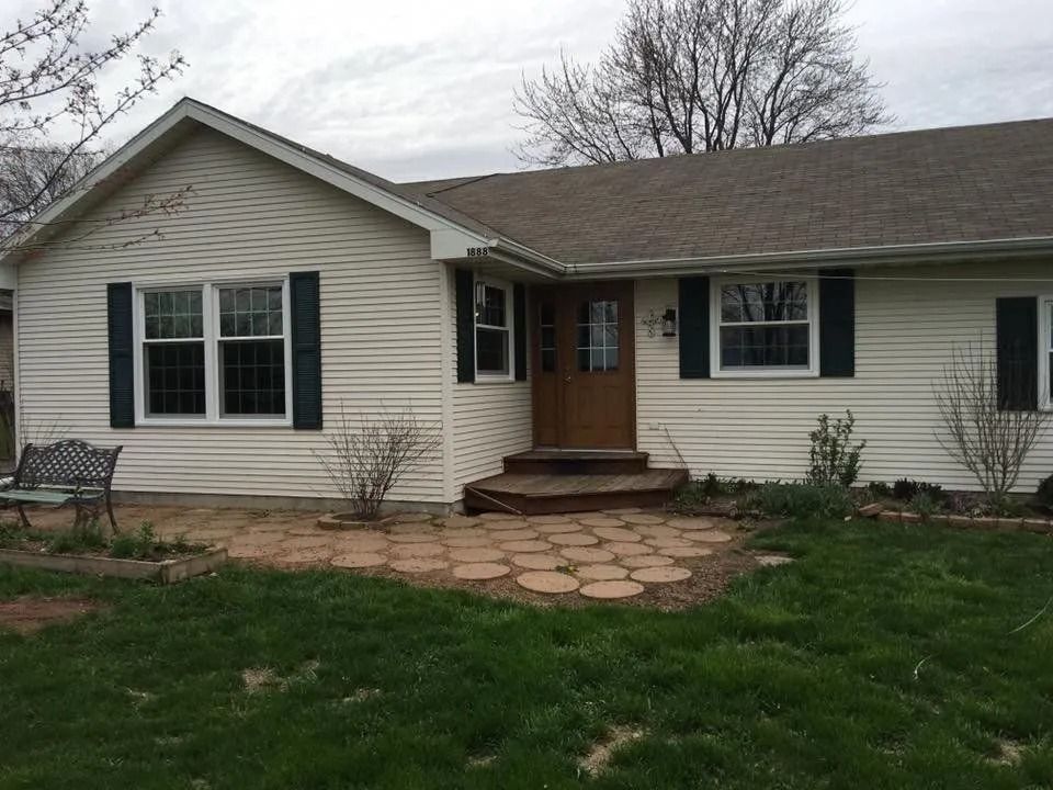 Beige house with a brown door, dark shutters, and a stone patio. Green lawn and a cloudy sky.