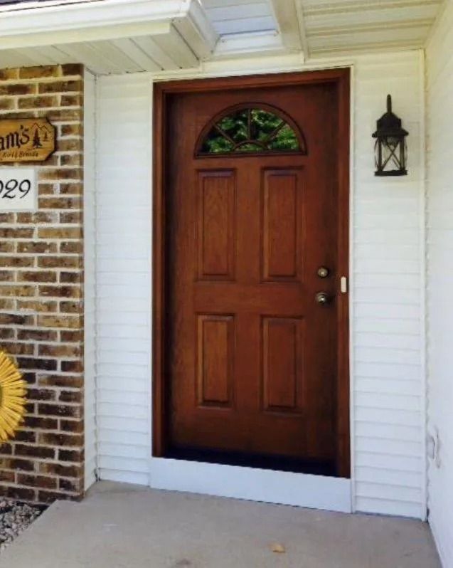 Wooden front door with arched window, flanked by white siding and brick.