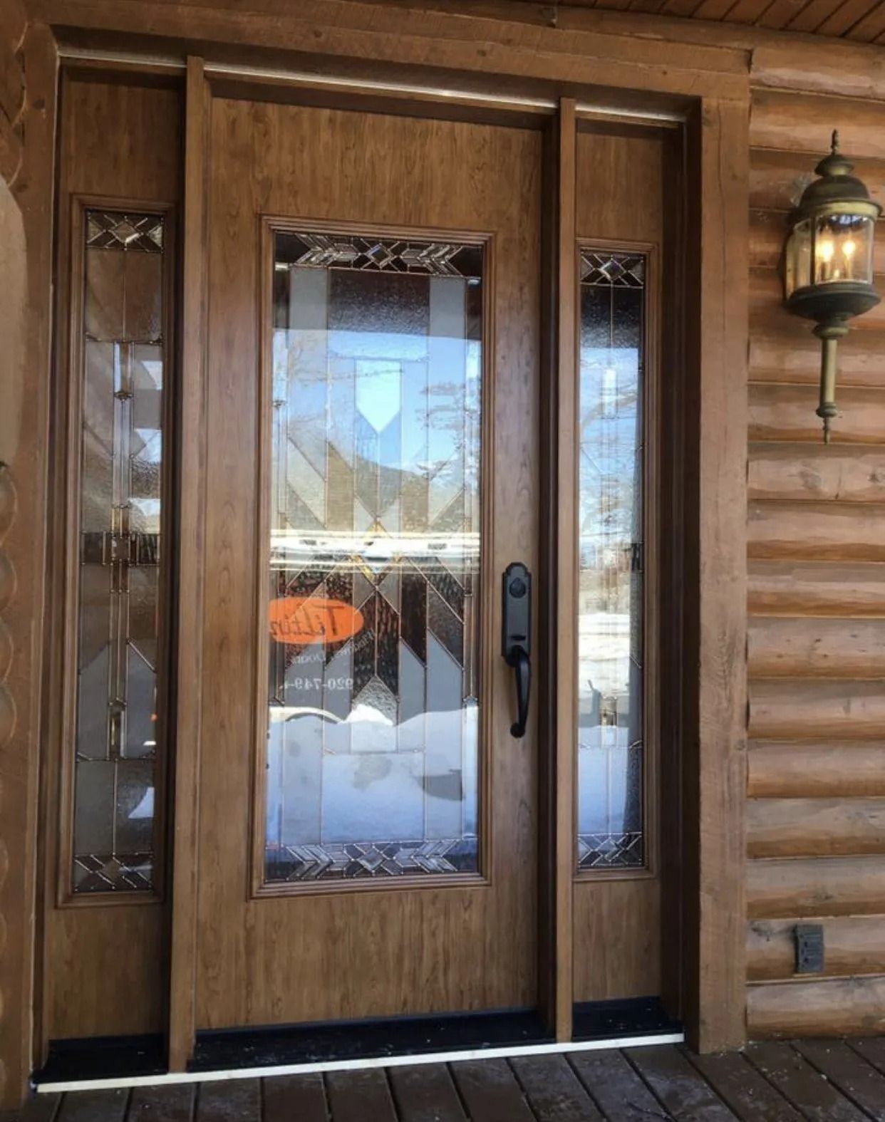 Wooden front door with glass panels and sidelights on a log cabin porch, with a brass lantern.