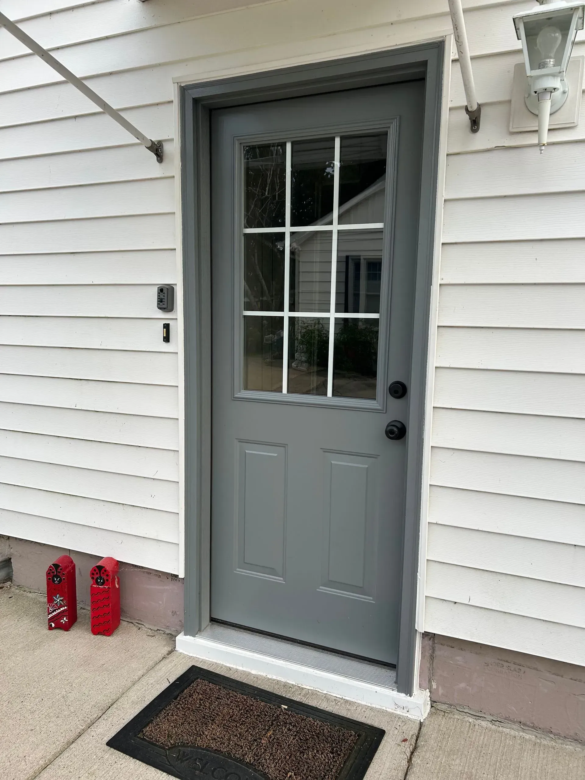 Gray door with glass window and black handle, set in a white-sided building; a welcome mat is at the threshold.