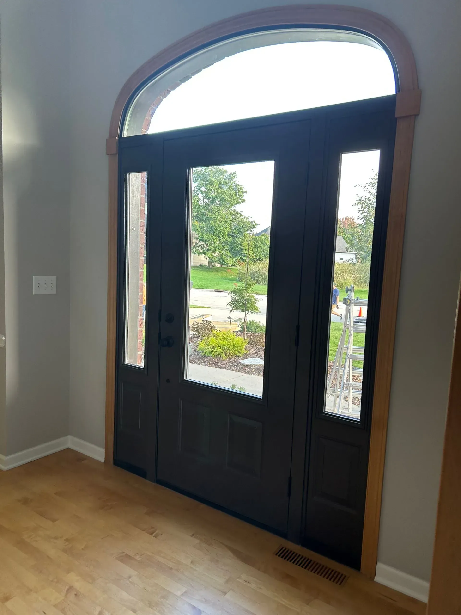 Entryway with black front doors and arched window above, set in a light-colored interior with wooden floor.