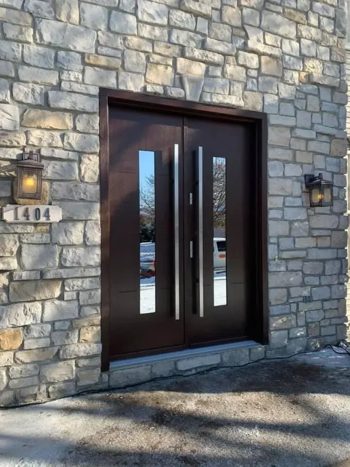 Double brown doors with vertical glass panels and silver handles in a stone building.