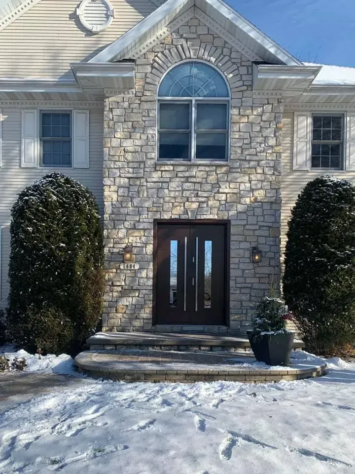 House exterior with stone facade, dark wood door, two evergreen shrubs, snow on ground.