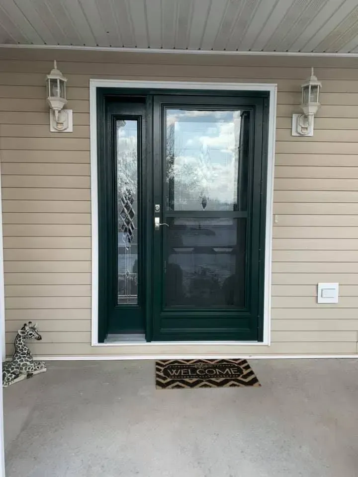 Dark green front door with sidelight, welcome mat, beige siding, white trim and sconces.