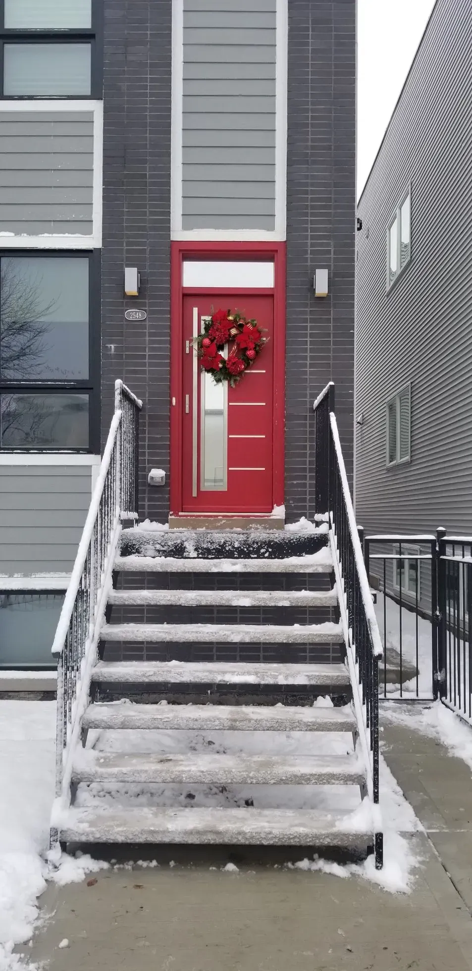 Snowy front steps leading to a modern red door with a wreath.