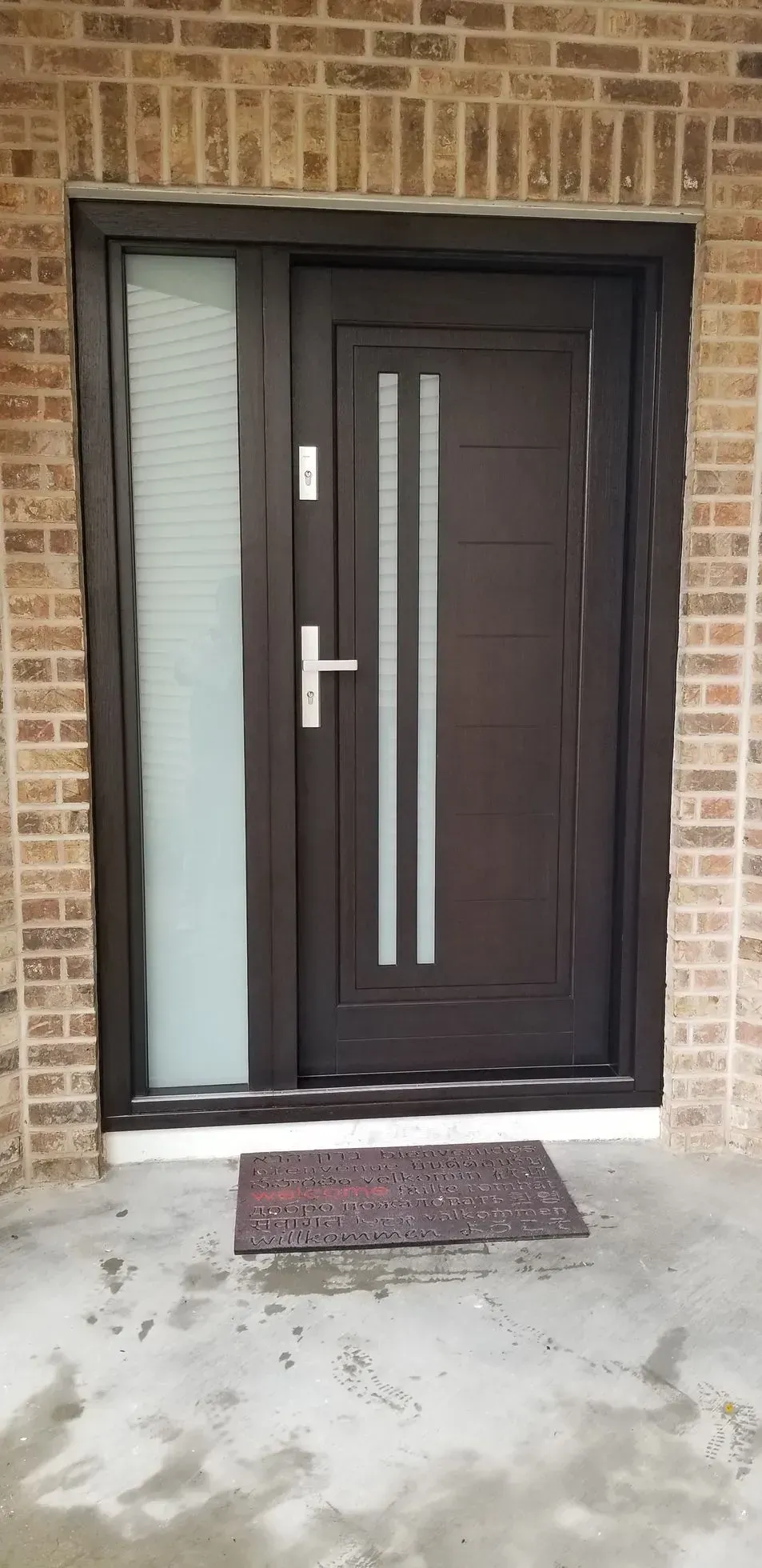 Brown door with sidelight, brick wall, and a welcome mat.