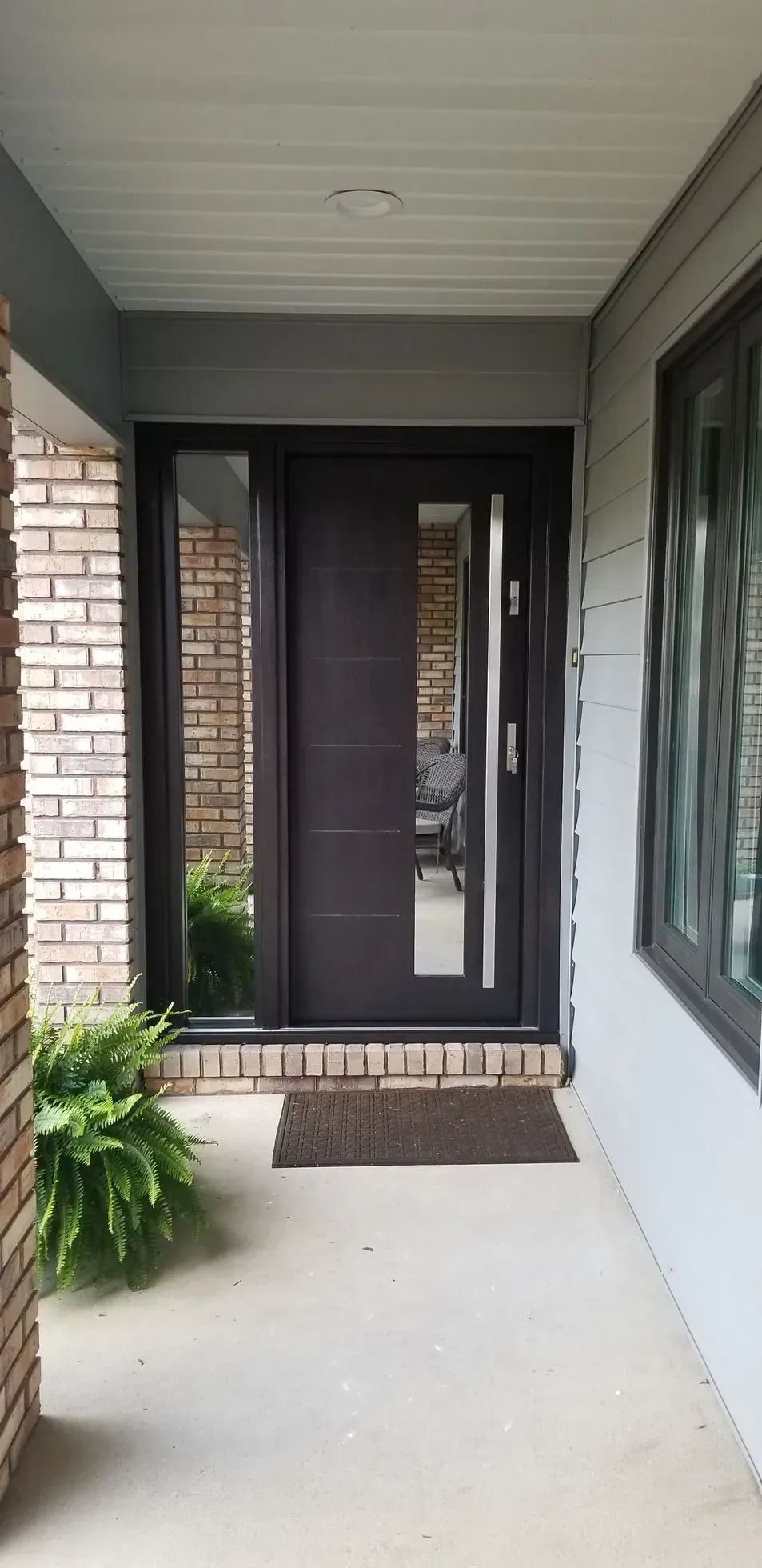 A dark brown front door with a side window and a welcome mat on a porch. Brick and light gray walls.