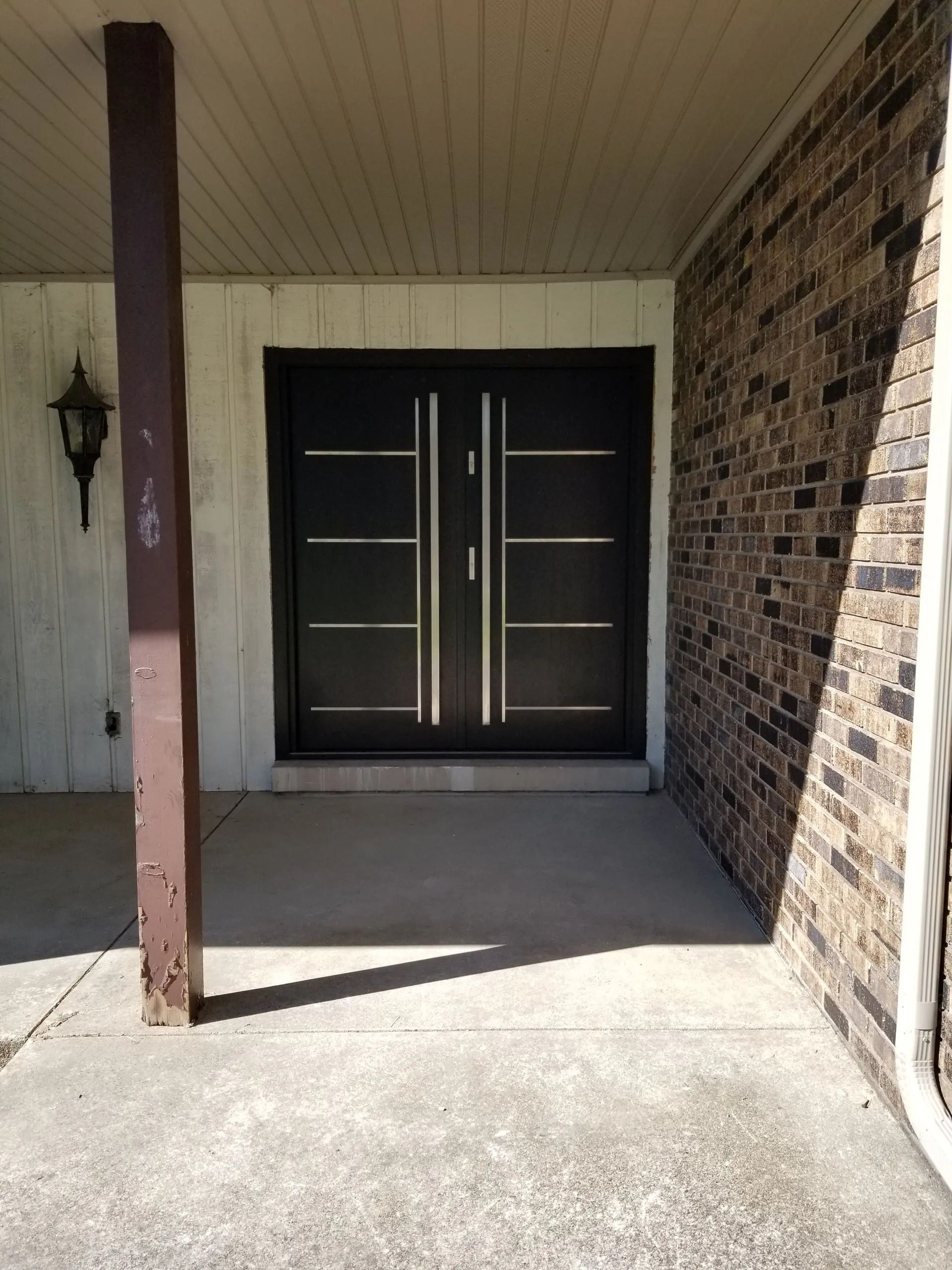 Black double doors with silver handles on a concrete porch, framed by a brick wall and a brown support beam.