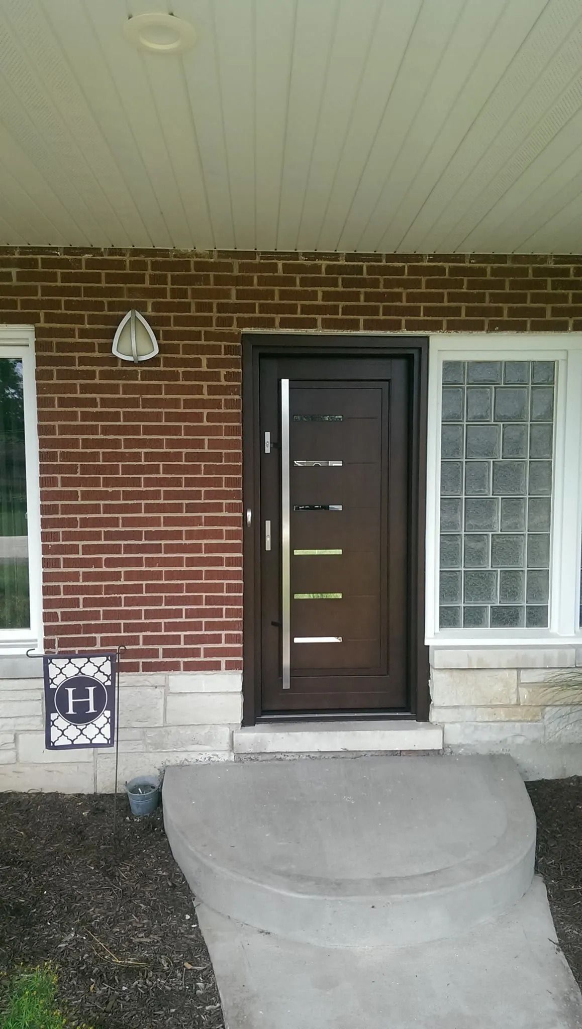 Brown front door with vertical silver handle, brick wall, concrete steps, and a side window.