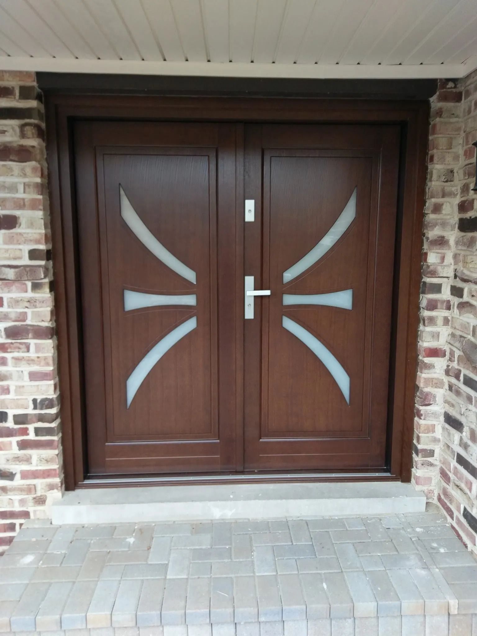 Double brown wooden doors with decorative frosted glass, silver handle, and brick surround.