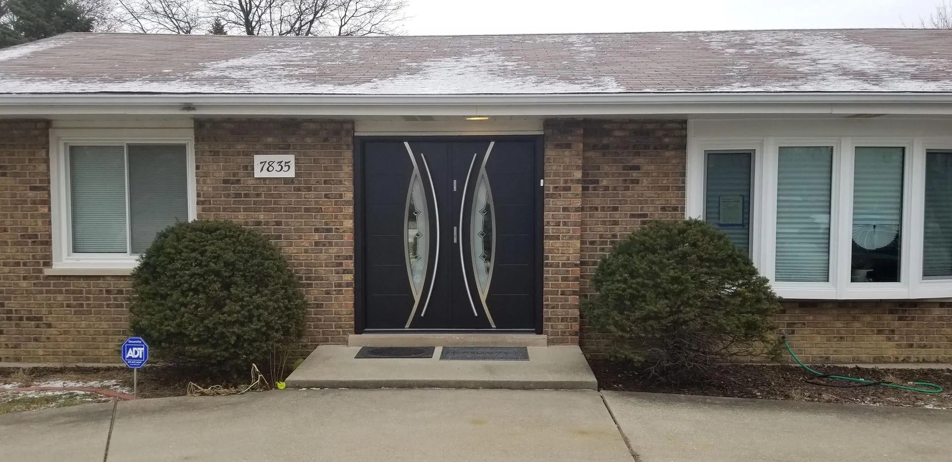 A brick house with black double doors, windows, and two green bushes flanking the entrance.