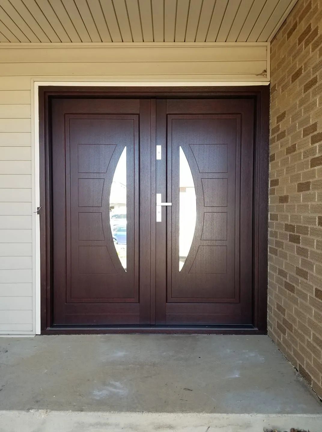 Double brown front doors with oval glass panels, leading to a building with brick and siding.