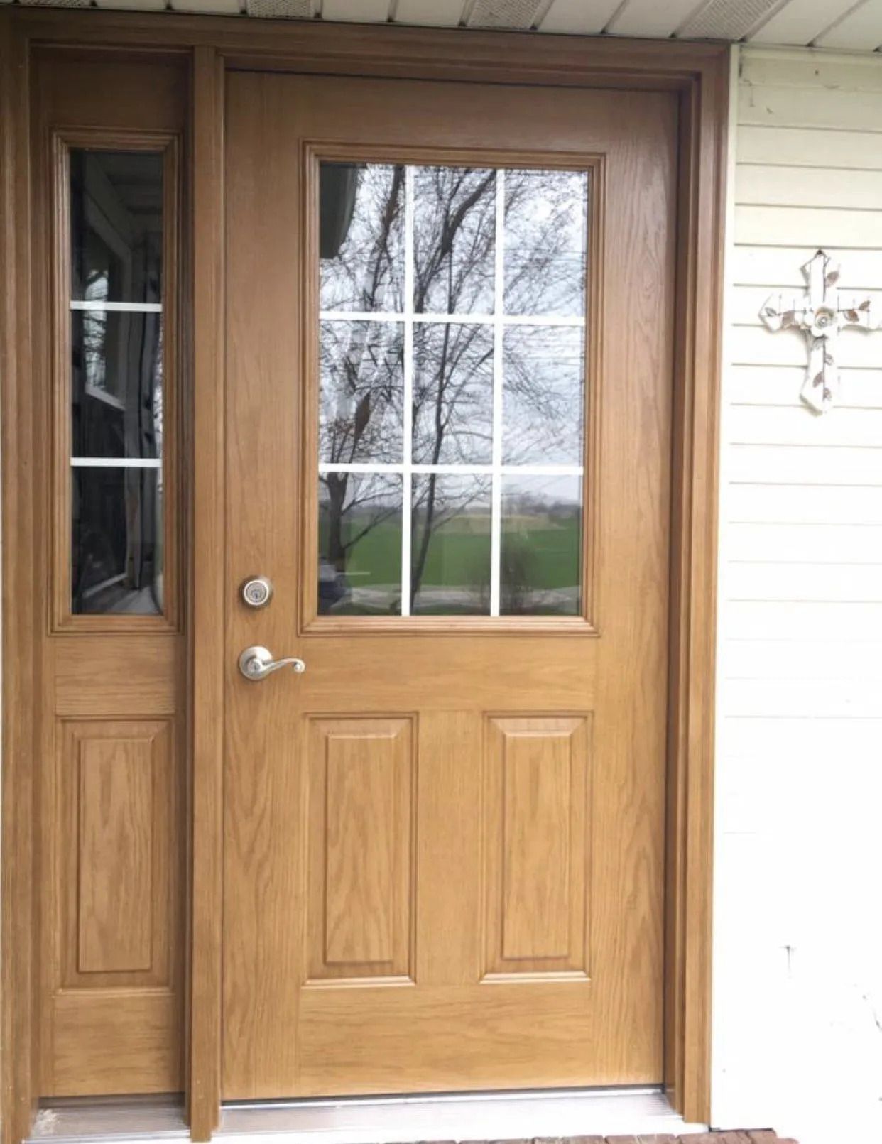 Wooden front door with glass window and sidelight. Tan siding. White cross decor on the wall.