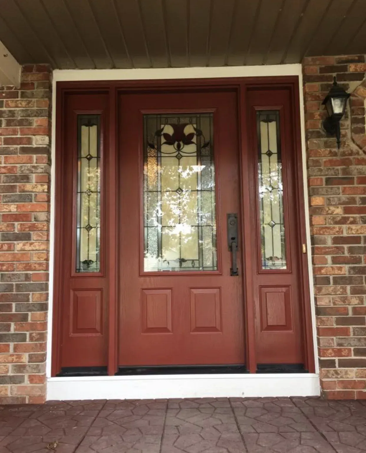 Red front door with stained glass, sidelights, and black hardware in a brick building.