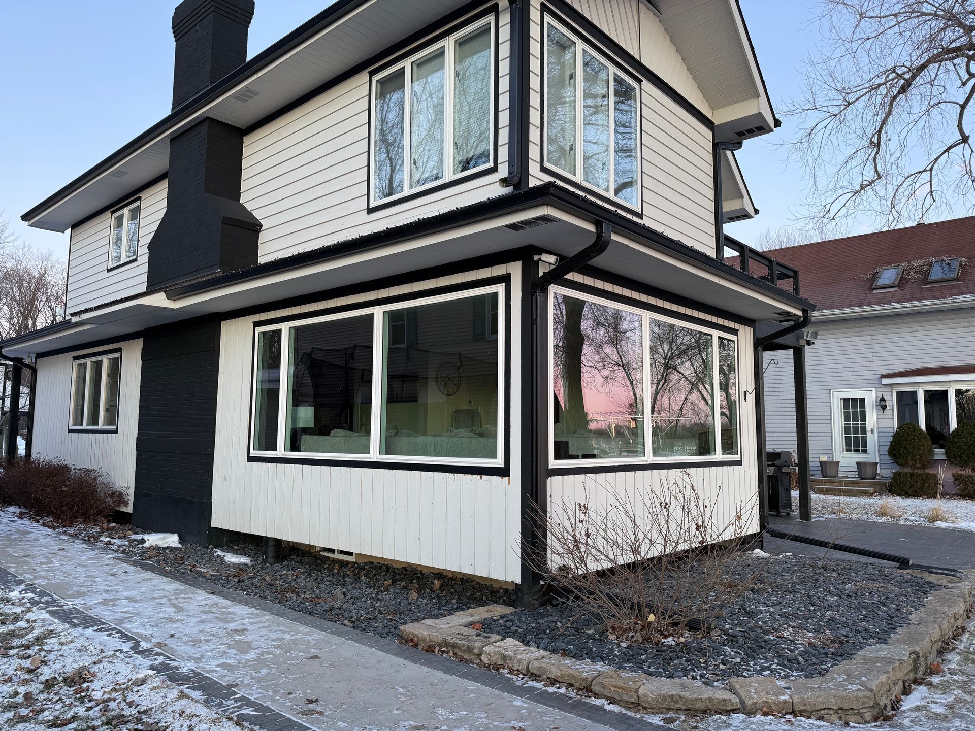 Two-story white house with black trim, chimney, and large windows. Winter scene with snow and blue sky.