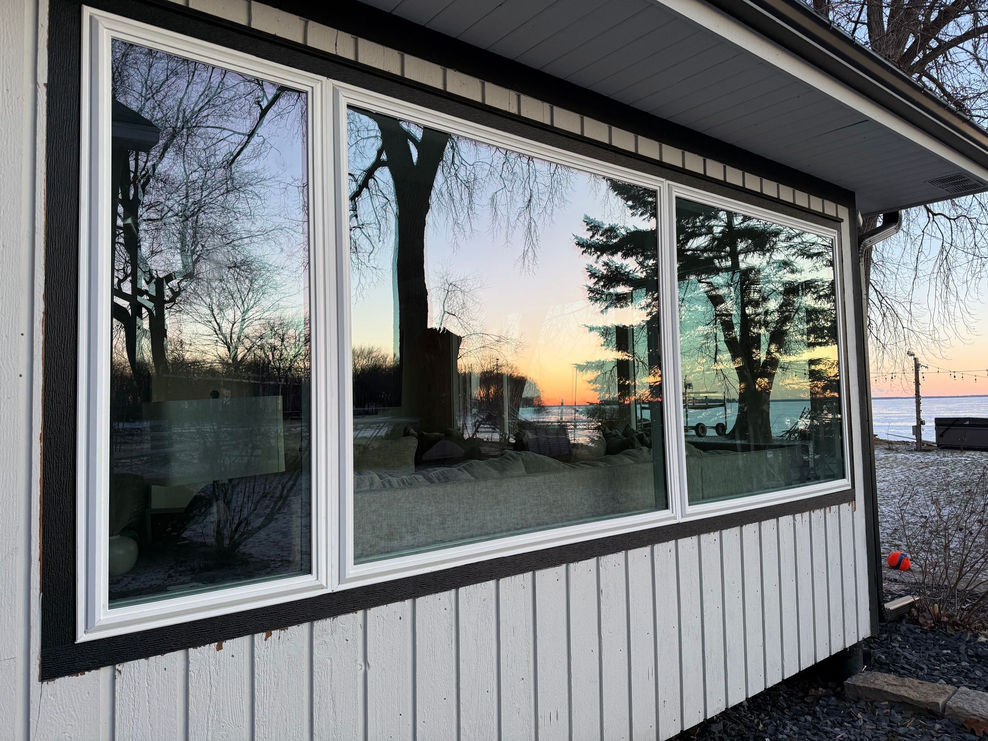 Large windows reflecting a colorful sunset and trees; mounted on a white and dark-trimmed building near water.