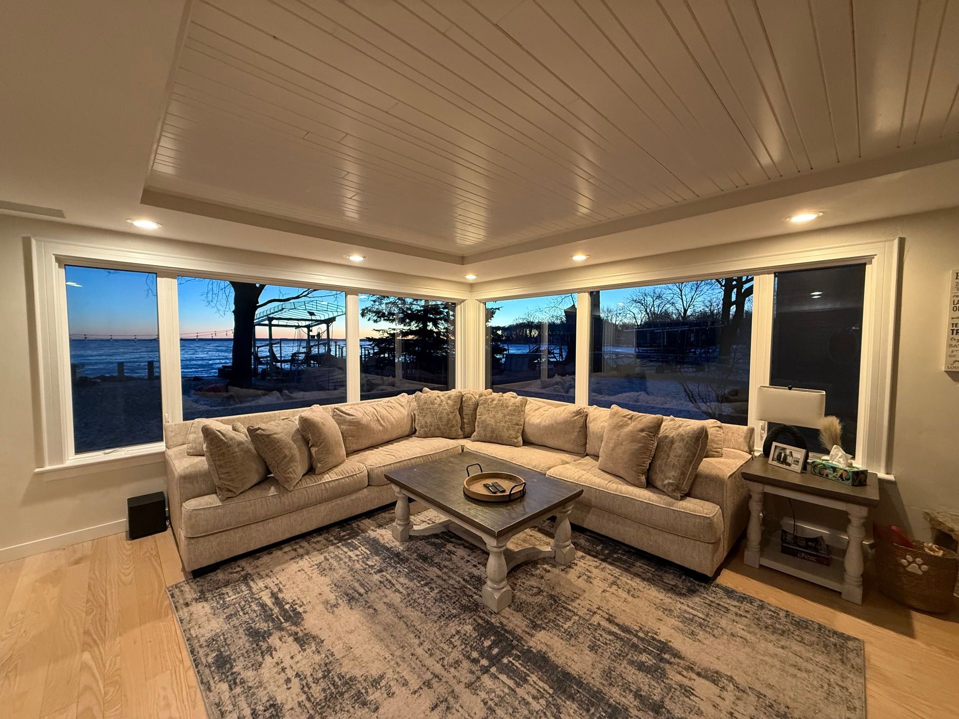 Cozy living room with sectional sofa, rug, coffee table, and large windows overlooking water at dusk.
