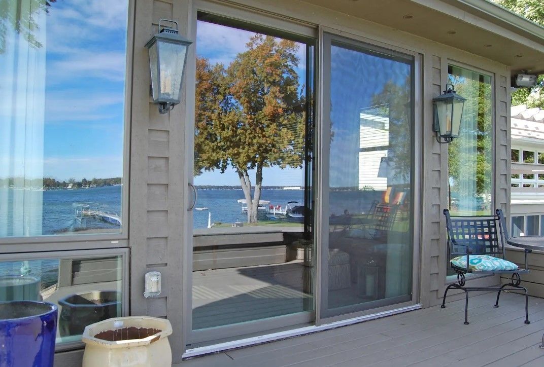 Sliding glass door and windows reflecting a lake scene, outdoor deck with a chair, and wall lanterns.
