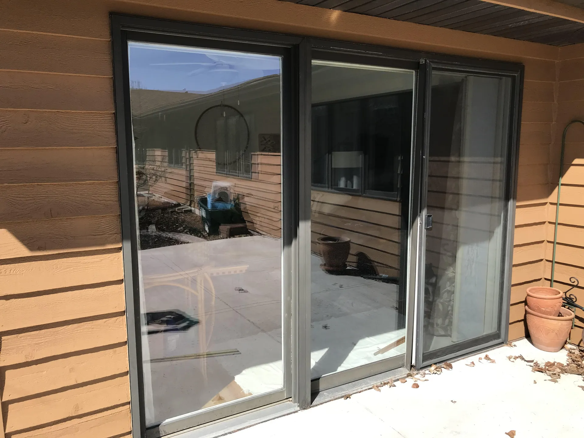 Sliding glass door, gray frame, reflecting outdoor patio and yard. Brown brick siding, sunny day.