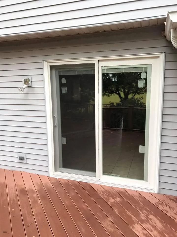 Sliding glass door on a wooden deck. Light gray siding and white door frame. Green trees visible through the glass.