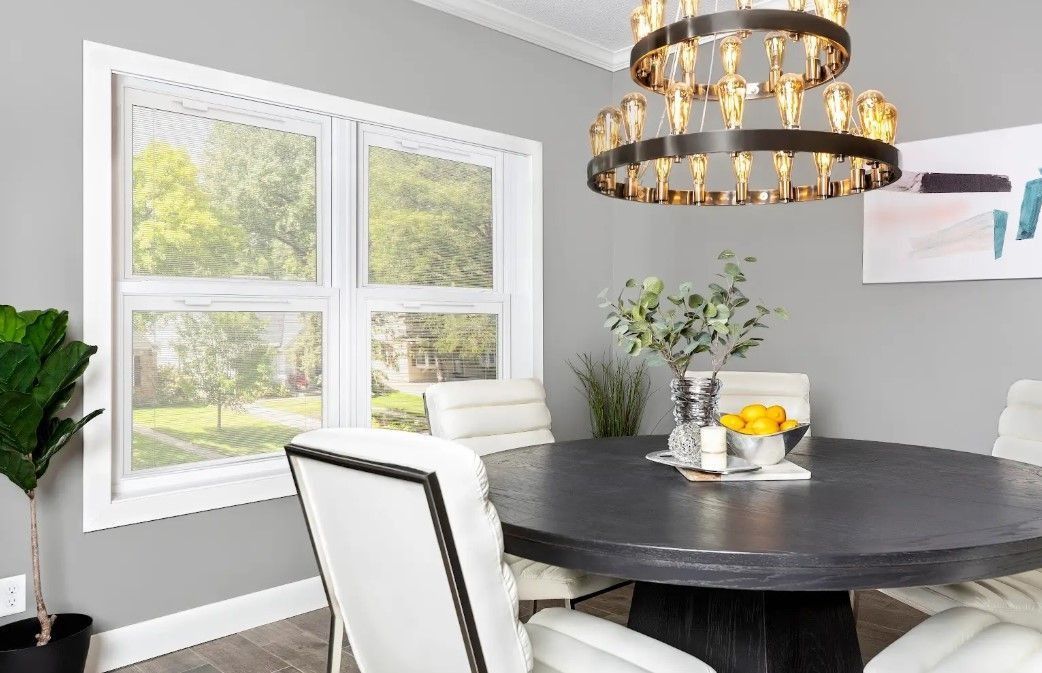 Dining room with round black table, white chairs, large window, chandelier, and artwork on gray wall.