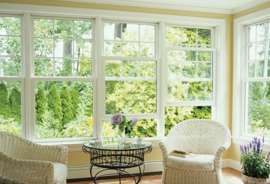 Sunroom with white-framed windows overlooking lush green trees. Two wicker chairs and a glass-topped table inside.