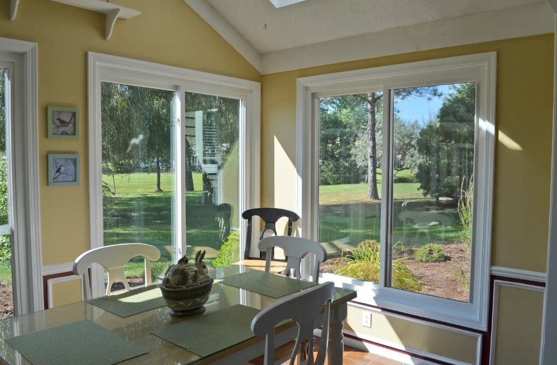 Dining room with a table, chairs, and large windows overlooking a green yard and trees.