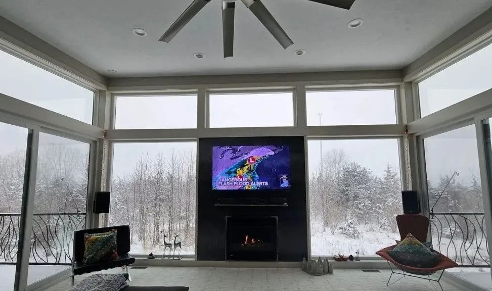 Living room with snowy view, large windows, fireplace, TV, and ceiling fan.