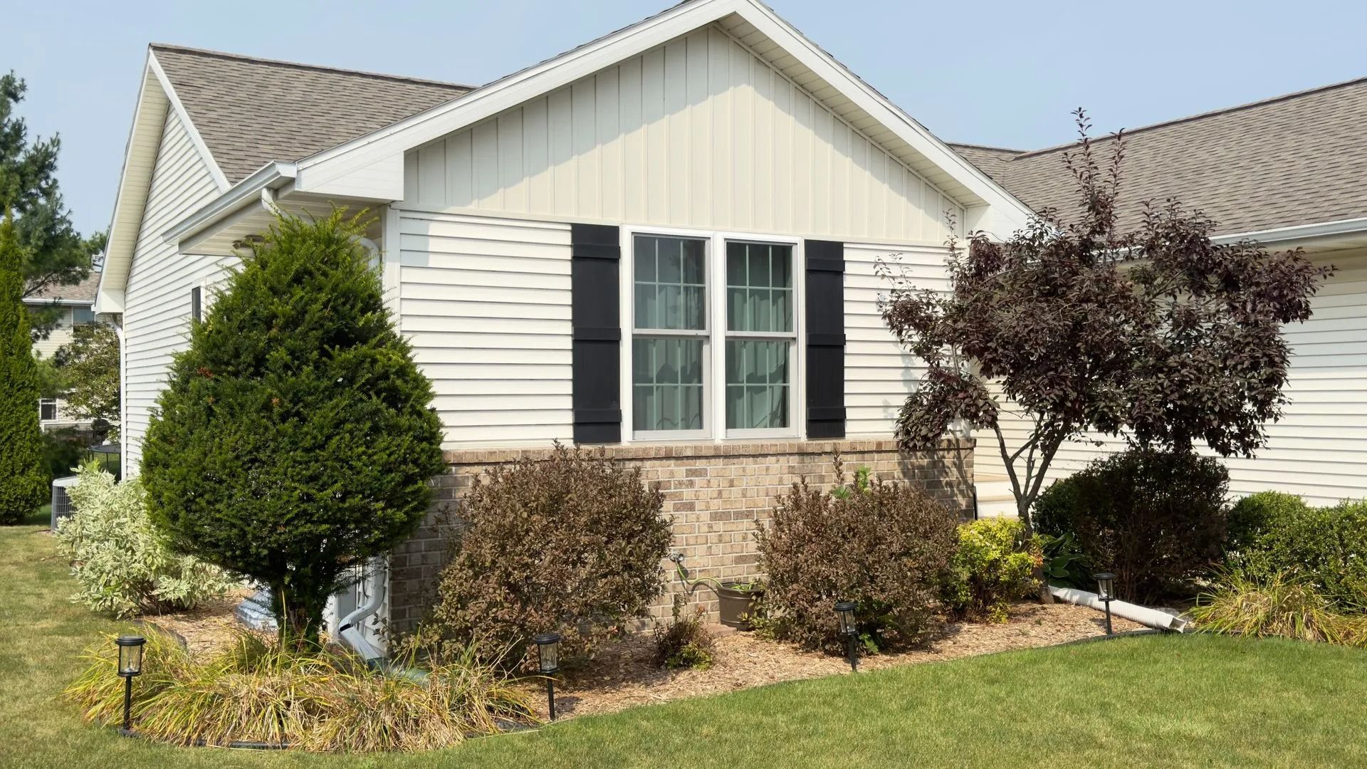 Tan and white house with black shutters, green and brown landscaping, and a blue sky.