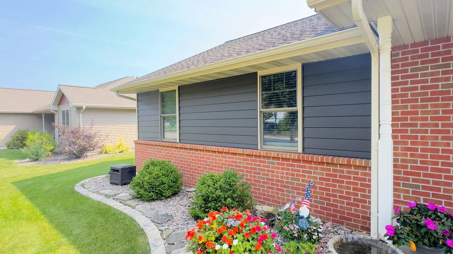 Side view of a house with brick and dark gray siding, featuring windows, shutters, and landscaping.
